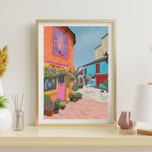 A framed art print depicting a colorful street scene with orange and blue buildings, displayed on a light wood shelf next to decorative items.