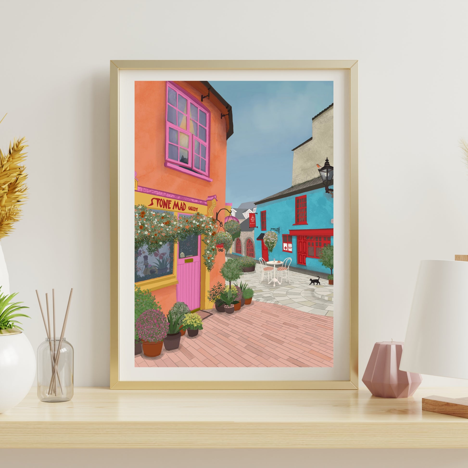 A framed art print depicting a colorful street scene with orange and blue buildings, displayed on a light wood shelf next to decorative items.