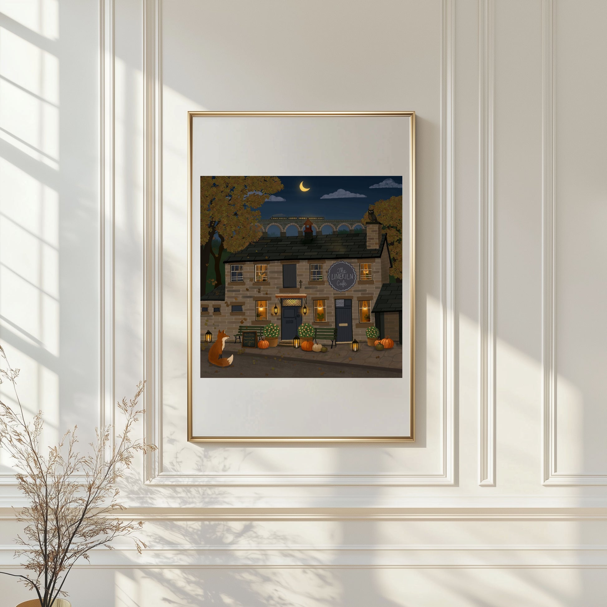 A framed art print depicting a cozy autumn night scene with a stone cottage, pumpkins, and a full moon, displayed on a white paneled wall with soft window shadows.
